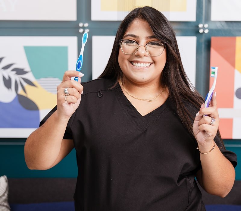 A woman in a glasses is smiling and holding two toothbrushes in her hands